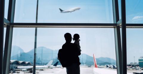 Father carrying daughter, looking out of airport window at a plane taking off
