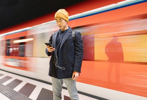 Asian man wearing a cap looking at his smartphone in front of a moving train.