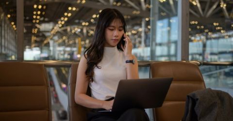 Asian woman at airport talking on mobile phone and with a laptop in her lap