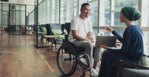 A person using a wheelchair and another person with a laptop are engaged in a conversation in a modern, sunlit office space with large windows and comfortable seating.