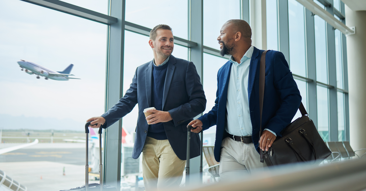 Two businessmen walking with suitcase in an airport with an airplane taking off outside 