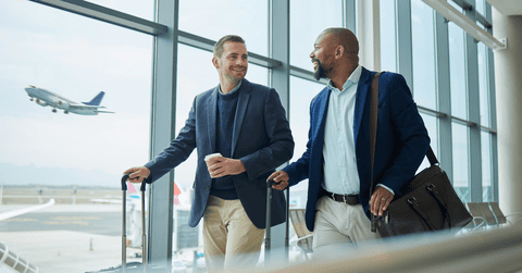 Two businessmen walking with suitcase in an airport with an airplane taking off outside 