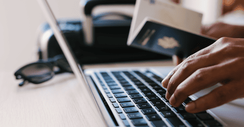 Close-up of man's hand typing in laptop keyboard, with passport in hand, sunglasses and briefcase alongside